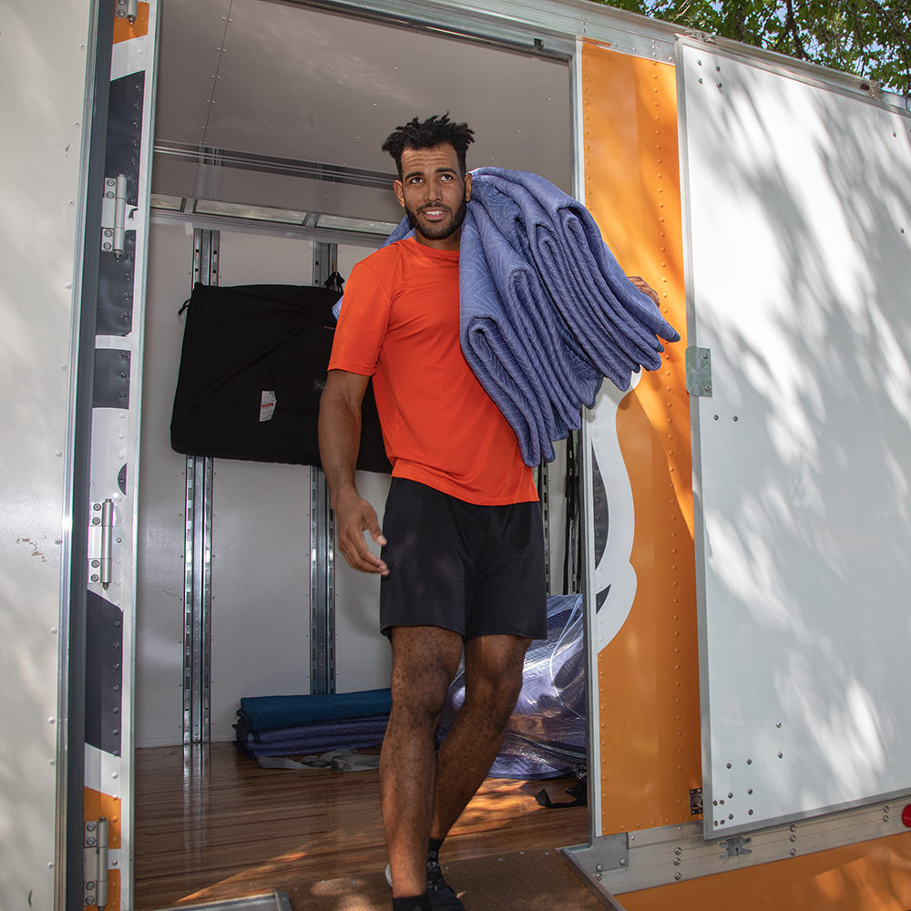 An Einstein mover stands in the doorway of a moving truck. He has moving pads slung over his shoulder