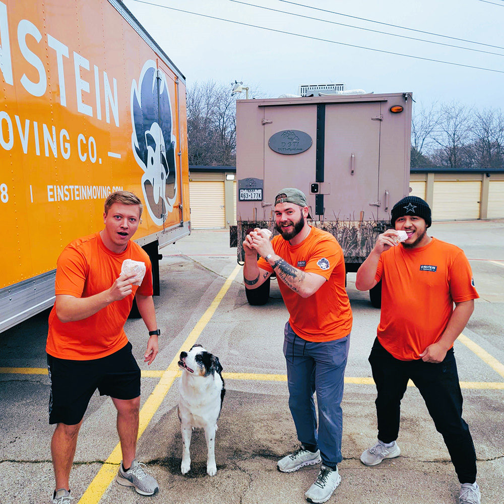 Einstein movers stand with sandwiches in front of their moving trucks, and a dog!