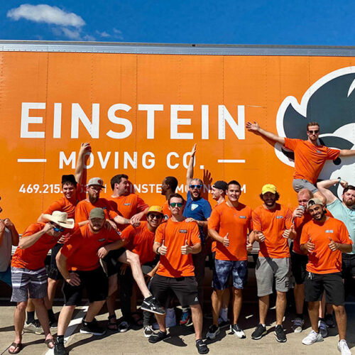 Group of Einstein Moving Company team members in orange shirts posing energetically in front of a branded moving truck.