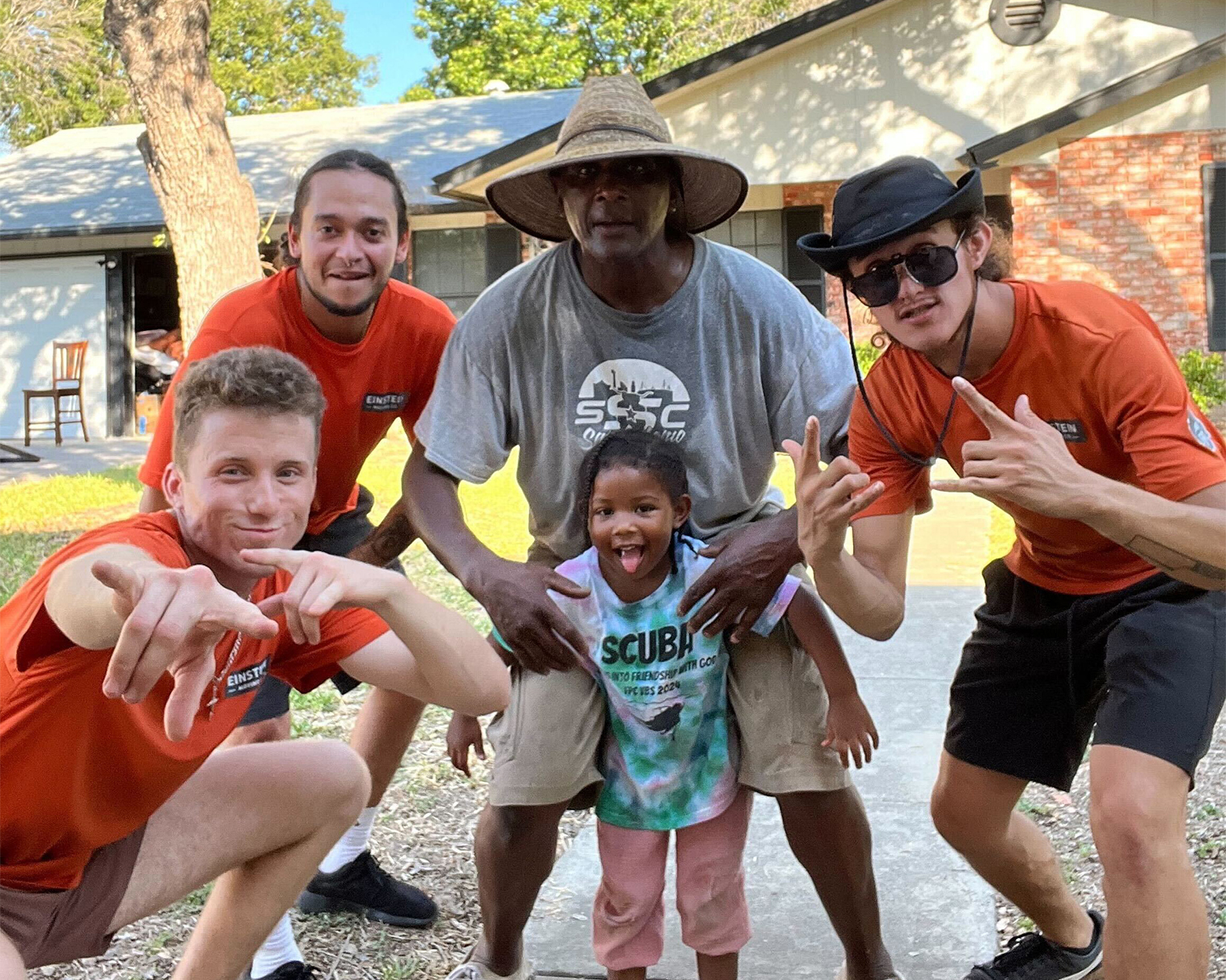 A cheerful group of Einstein Moving Company team members posing with a father and his daughter outside a home. The movers are wearing orange company shirts and crouching or pointing playfully at the camera. The little girl in the middle is smiling and sticking out her tongue, and the father stands protectively behind her, wearing a wide-brimmed hat.